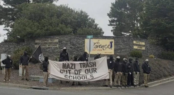 A group of Nazis stands on a corner holding a sign that says "Nazi trash out of our schools"