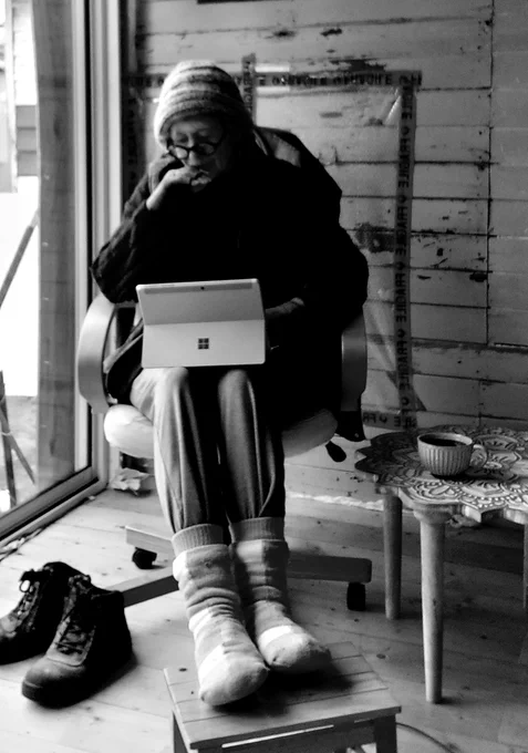 Woman in thick winter clothes sits inside by a large window, working on a laptop. Work boots and coffee cup are nearby. The wall behind her needs repair and painting