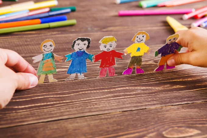 Foto: Auf einem Tisch ist eine Gruppe von ausgeschnittenen Vorlagen zu sehen. Diese sollen fünf Kinder darstellen, die sich an der Hand halten. Im Hintergrund sind Stifte zu sehen. 