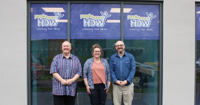 Three people smiling to camera standing in front of windows with decals showing the People Know How logo.