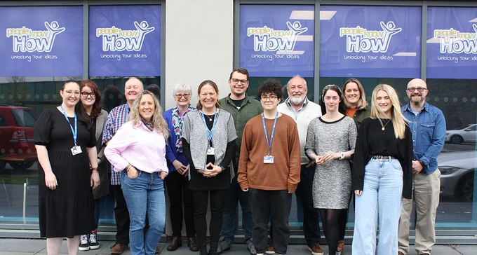 A group of staff and trustees from People Know How standing in front of windows with decals showing the People Know How logo. 
