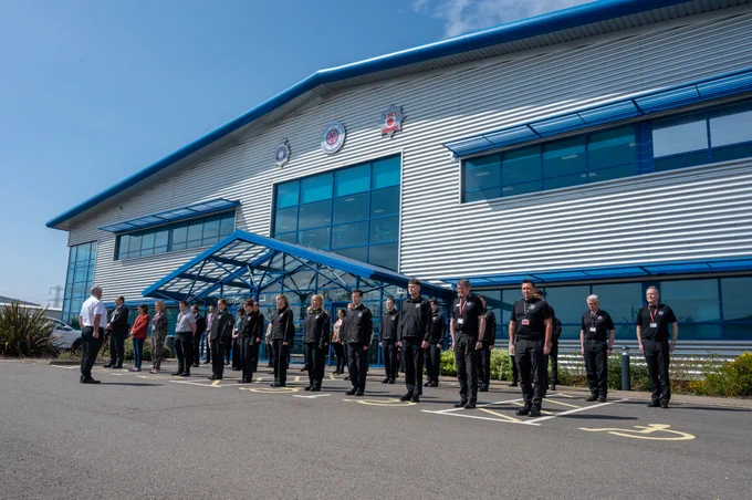 Firefighters and staff outside Service Headquarters building. Sky is blue and 3 emblems of Northants Fire, Police and OPFCC can seen.