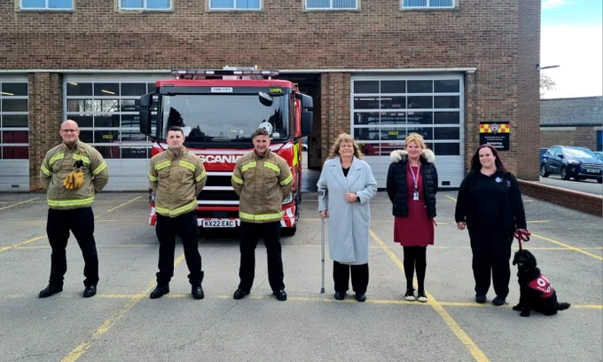 Kettering Fire Station with one of the engines outside. There are 3 firefighters from Red Watch standing in line alongside Tracey and Jo from Health and Safety and Ffion from IT along with Missy a wellbeing dog. 