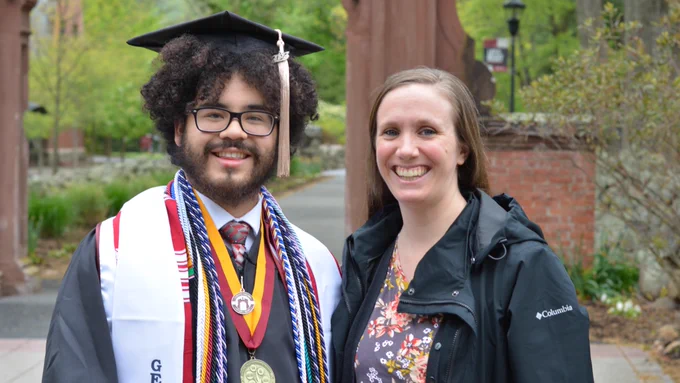 Paolo and Alexandra pose for a photo in front of the Ramapo College Arch, which is cut off at the top out of frame. Paolo is dressed in all regalia for his student speaker photos for the upcoming undergraduate commencement. He is on the left and Alexandra is on the right.