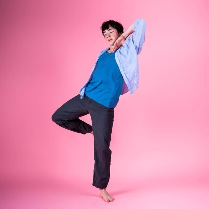 A young male dancer standing on one foot with his other leg bent dressed all in blue against a pink background.
