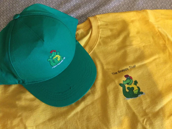 A green baseball cap and a bright yellow T-shirt lying on a table. Both items have the snappy croc logo on them.