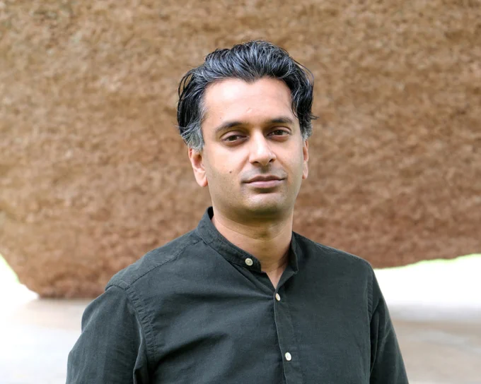 A South Asian man in a black shirt with wavy hair stands in front of a large stone.