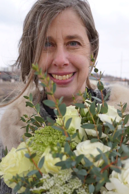A smiling white woman with light brown/dark blonde hair blowing in her face holds a bouquet of flowers outside.