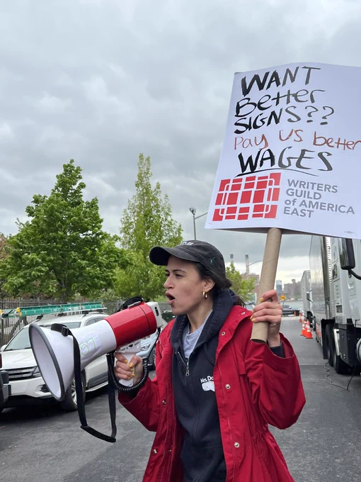 A person in a bright red jacket holding a bullhorn and holding a picket sign which says “Want better signs? Pay us better wages.”