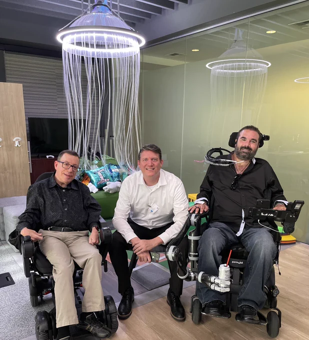 Image shows a brown hair male, middle aged in a power wheelchair beside a blonde haired gentleman in the middle beside a bearded brown hair gentleman in a power wheelchair.