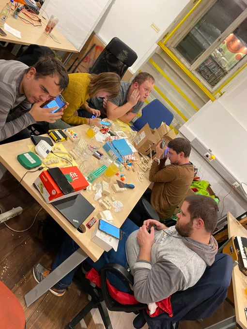 5 Menschen sitzen an einem Tisch und sind in ihre Arbeit mit Smartphones und Tastatur-Prototypen beschäftigt. Am Tisch befinden sich Tastaturen, Knetmasse, Schachteln, Papiere und Gläser.