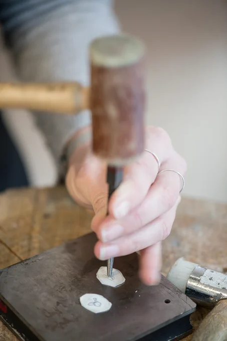 A hand holds a metal jewellery tool which is being hammered into two small pieces of cut silver, on a metal block. 