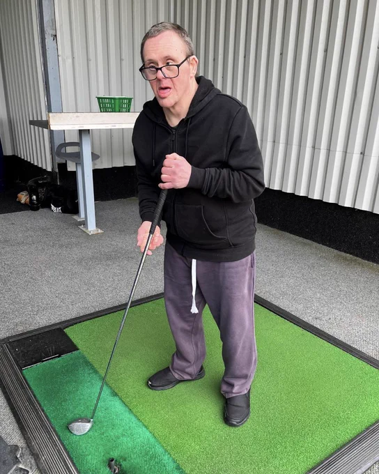 A person is stood on a golf mat ready getting ready to hit their ball. They're stood looking at the camera. 
