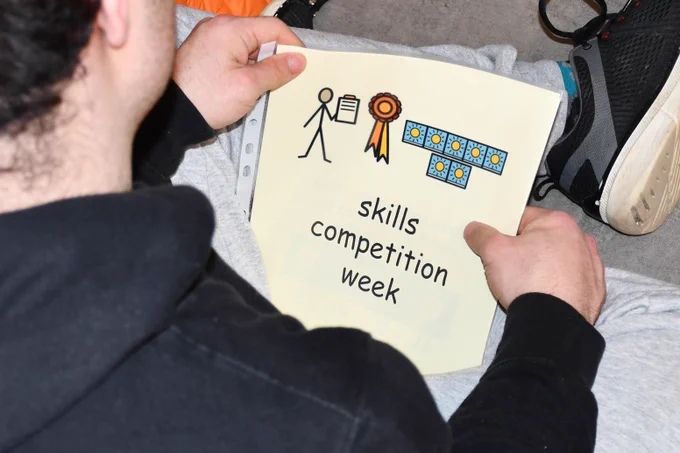 A young man in a black hoodie holding a sheet that says "skills competition week"