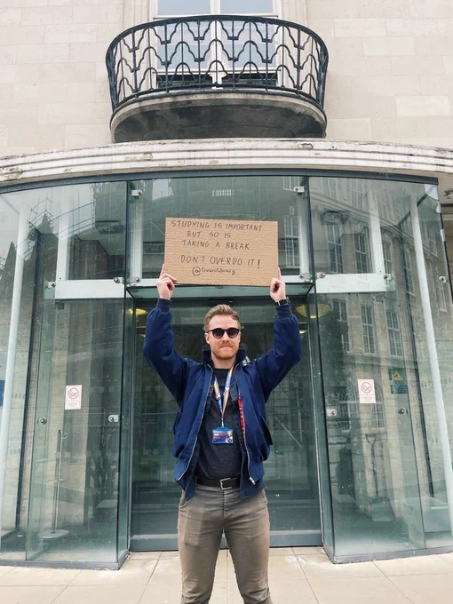 Library team member stands in front of the glass entrance doors of the Harold Cohen Library. He is wearing sunglasses and holding up a cardboard sign encouraging students to take a break from studying.
