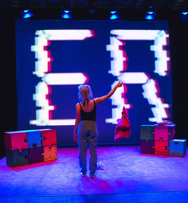 Alice stands facing a screen with error on it with her right arm stretched out having  just dropped a t shirt on the floor
