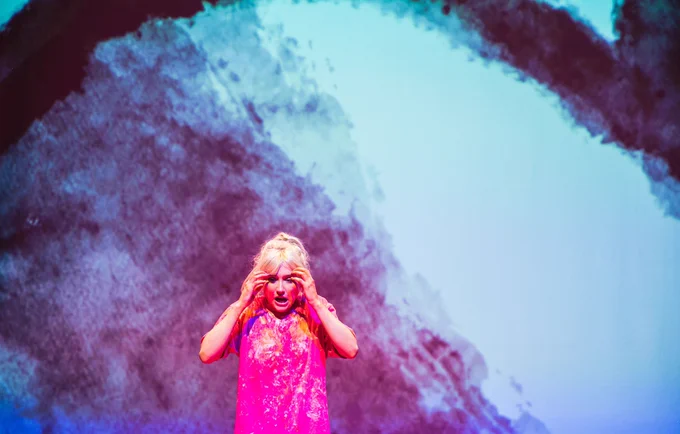 Alice stands with her hands to her head with a black and white screen behind her