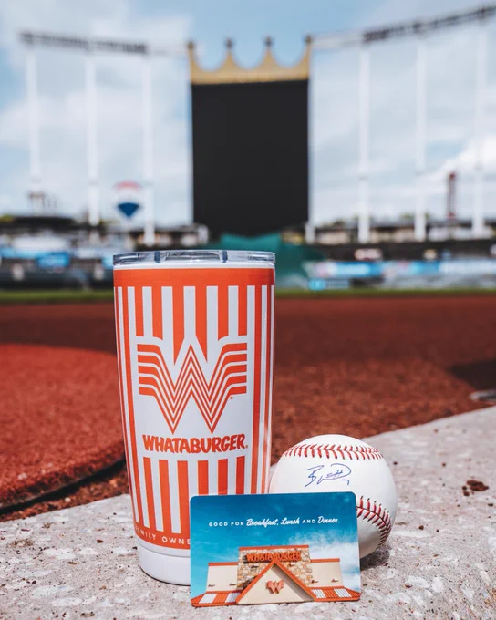 A photo of a $25 Whataburger gift card, a Whataburger-branded Yeti Rambler 20-ounce cup, and a baseball signed by Bobby Witt Jr. placed on the top step of the Royals dugout. Kauffman Stadium and the CrownVision video board can be seen in the background of the photo.