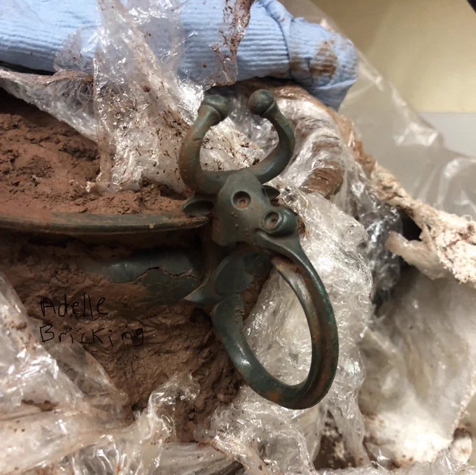 A copper alloy bowl with an adorable, cartoon-like decorative ox head escutcheon on the rim. The ox has curving horns with round caps on the ends, little ears that poke out the side of its head, large round hollow eyes and matching nostrils, with the handle protruding from its mount and curving back down to the bowl’s body. The bowl is still covered in mud and packed with cling film and blue tissue.
