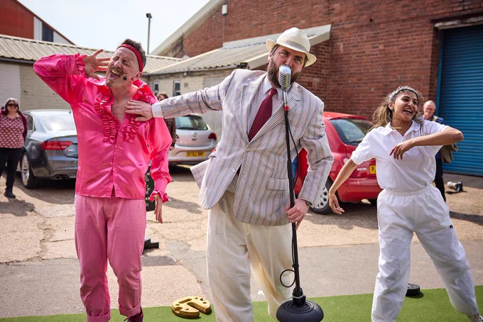 The three bowls perform a song & dance routine, man in middle holds an old fashioned microphone