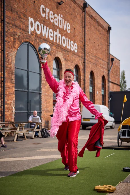 Man in garish costume & feather boa raises a Glitterball trophy, standing on green carpet