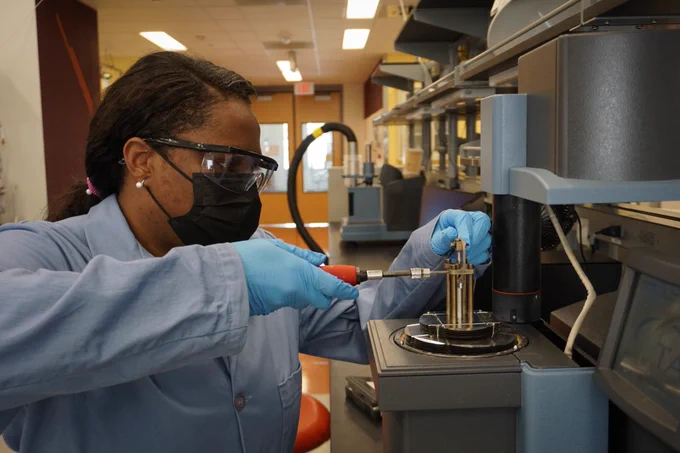 Tiffany Thompson wears safety glasses and a lab coat while using a tool to adjust a laboratory instrument.