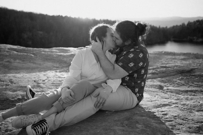 Black and white photo of a man and a woman sitting near a lake kissing at sunset.
