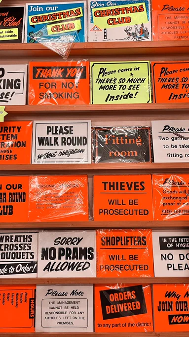 A selection of screen printed signs from Clarks brothers. There are  six shelves filled with signs printed on fluorescent orange, white or fluorescent yellow paper. They have black type showing a range of phrases, for example, “thieves will be prosecuted” “please walk around without obligation”