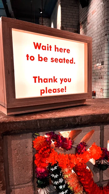 An illuminated sign with red text that reads “wait here to be seated. Thanks you please!”