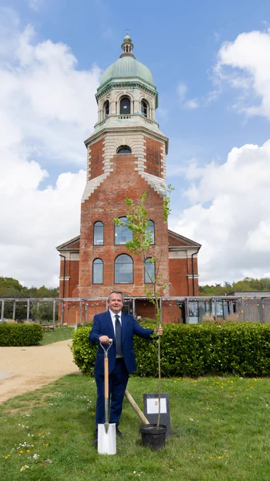 Cllr Humby with the tree and a shovel, standing where he planted the tree in the ground in front of the chapel at Royal Victoria Country Park.