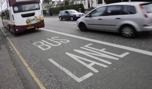 Photograph of a bus lane with the words 'Bus Lane' written on it and cars in the background and a bus on the lane