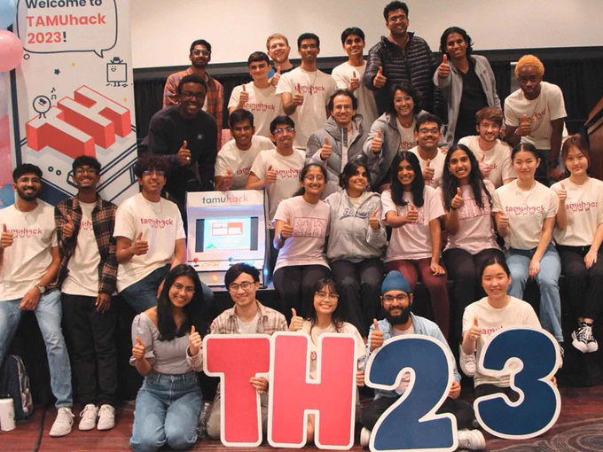 A group of TAMUhack students sitting on and in front of a stage doing a thumbs up.