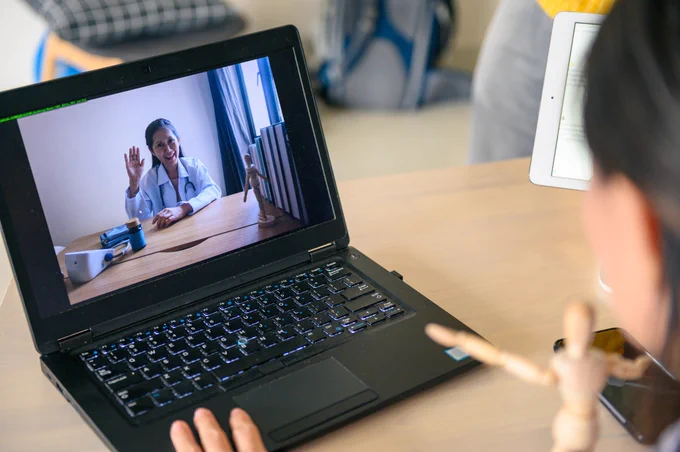 A doctor wearing a white coat and a stethoscope around her neck appears on the screen of a computer during a video conference with another individual. 