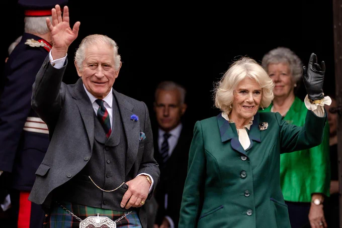 King Charles on the left wearing a grey suit and green red and blue kilt. He is waving and smiling. Queen camilla stands next to him on the right wearing a green blazer with a blue and green collar and black gloves. She is smiling and waving. A man and woman stand behind them.