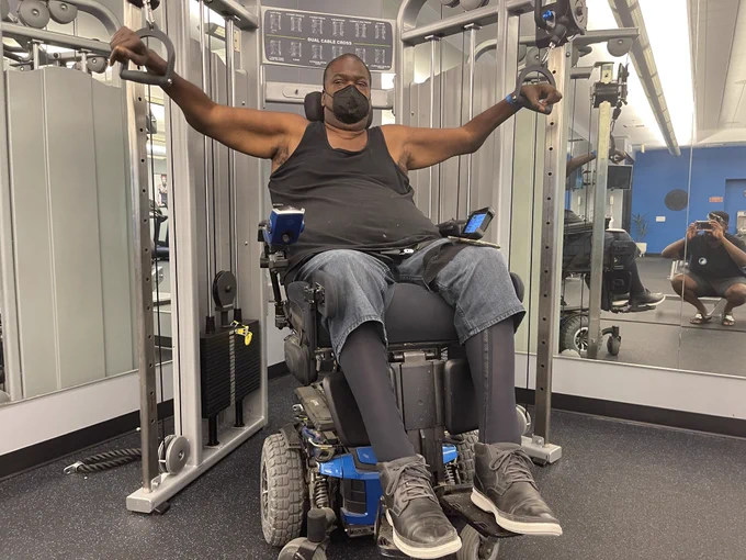 Image of Paul using cable machines while he is sitting in his Quantum Rehab power wheelchair. His arms are in the raised position in this photo and he is wearing a mask.