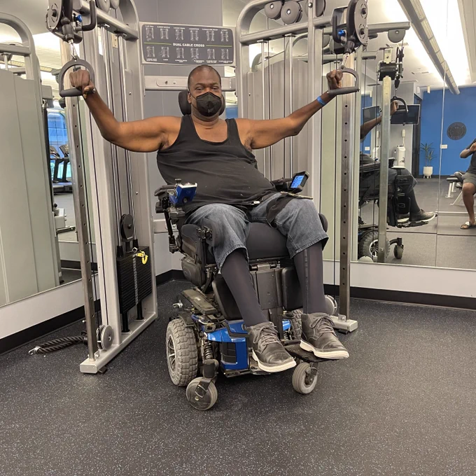A second image of Paul using cable machines while he is sitting in his Quantum Rehab power wheelchair. His arms are in the raised position in this photo and he is wearing a mask.