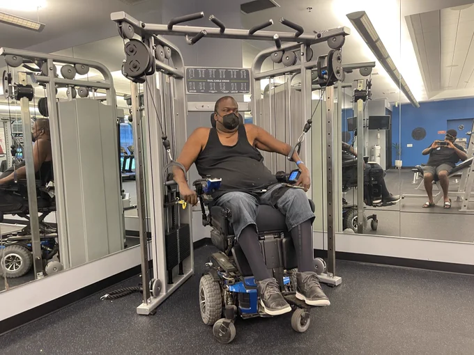 Image of Paul using cable machines while he is sitting in his Quantum Rehab power wheelchair. His arms are in the lowered position in this photo and he is wearing a mask.