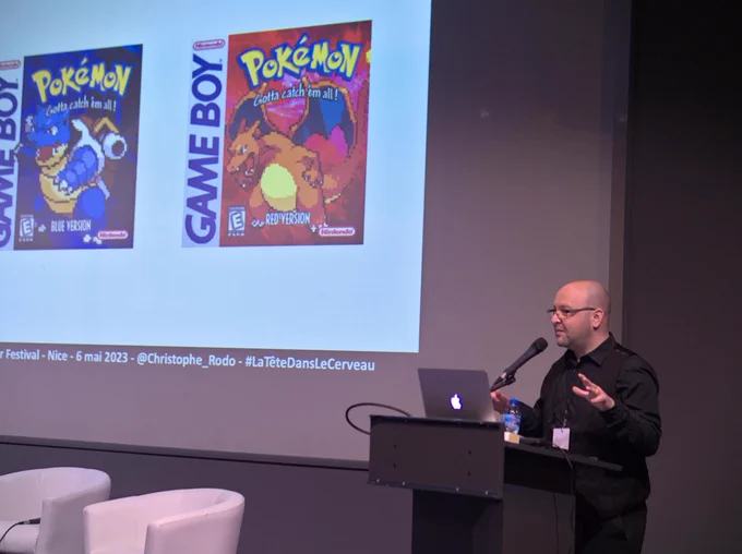 Christophe Rodo au premier plan, au pupitre d'une salle de conférence. À l'arrière plan, une slide avec les jaquettes de Pokémon Bleu et Pokémon Rouge, sur GameBoy.