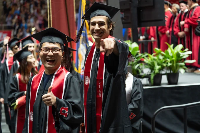 Photo of two graduates in regalia. One is giving the thumbs up sign, another pointing to the camera.