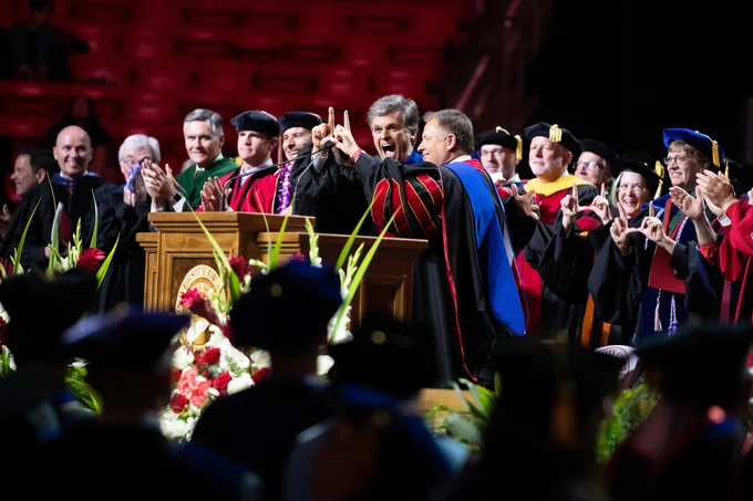Photo of the "platform" group standing up and clapping. President Randall and keynote speaker Tim Shriver are in the center joining in on a "Flash the U" with their hands. 