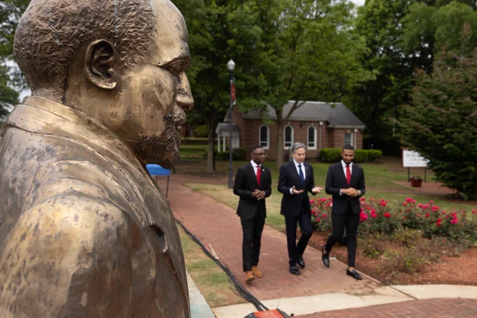 Secretary Blinken converses with two Pickering Fellows as they give him a walking tour of a university campus. In the foreground stands the golden bust of one of the university’s famous alumni.