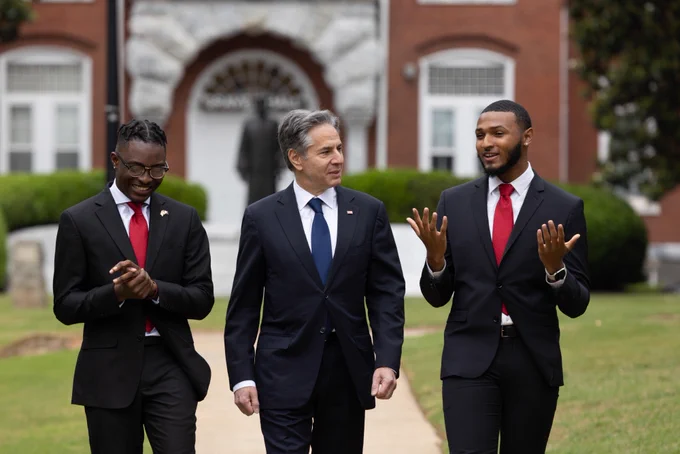 Secretary Blinken converses with two Pickering Fellows as they give him a walking tour of a university campus. In the background stands a bronze statue and a brick building with an arched entry.