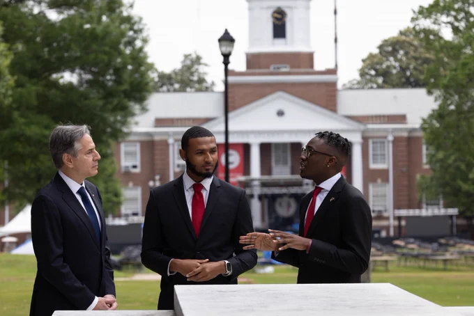 Secretary Blinken listens to two Pickering Fellows during a stop on a university campus tour. In the background stands a large brick building with white columns.