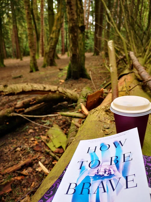 Image shows the book How To Be Brave by Louise Beech and a takeaway coffee cup on a fallen tree trunk in the midst of a pine wood, with pine trees in the background.