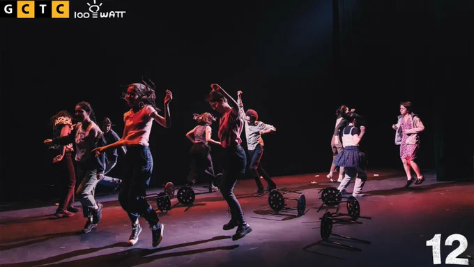 [Image Description: Text reads: "G C T C 100 Watt 12" Photo of eleven youth running in various directions around a stack of chairs spotlighted under a wash of red lighting.]