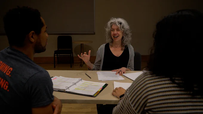 Gwendolyn Schwinke sits at a table, gesturing to two people whose backs are to the camera. 
