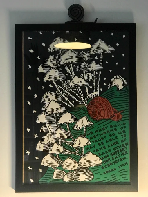 Photo of an enframed artwork by Justseeds with a snail on the grass, surrounded by a multitude of mushrooms. Under the snail, it reads: 'we must build networks of trust so we may be able to take care of each other and support our entire ecosystem' - Adaku Utah

On top of the artwork is another snail, made of metal.