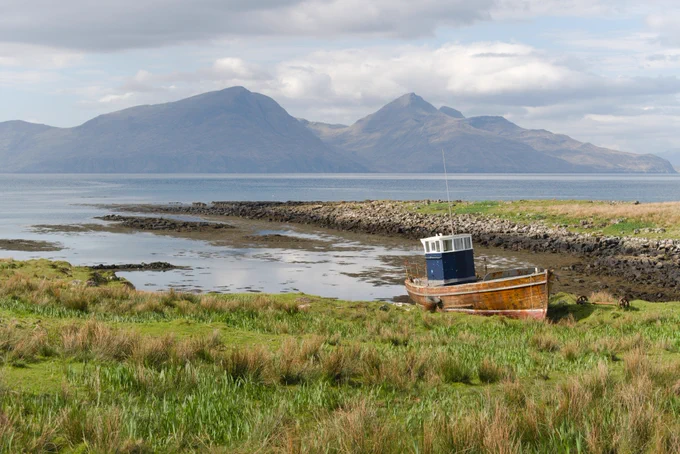 a wooden boat on grassy shoreline with a mountainous island in the distance 