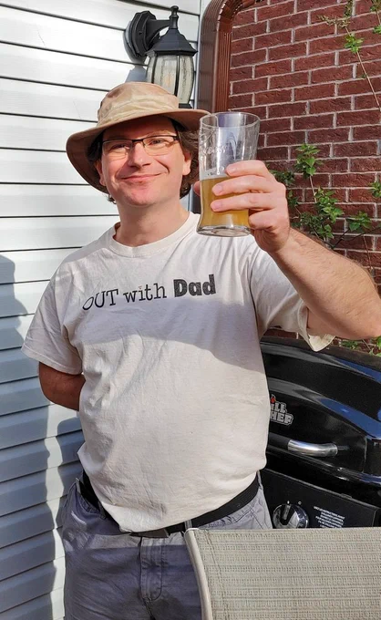 Greater and director Jason leaver standing outside in front of a brick wall hoisting a half drunk pint of beer in salute, smiling but be typically and wearing a white T-shirt with the Out with Dad logo.