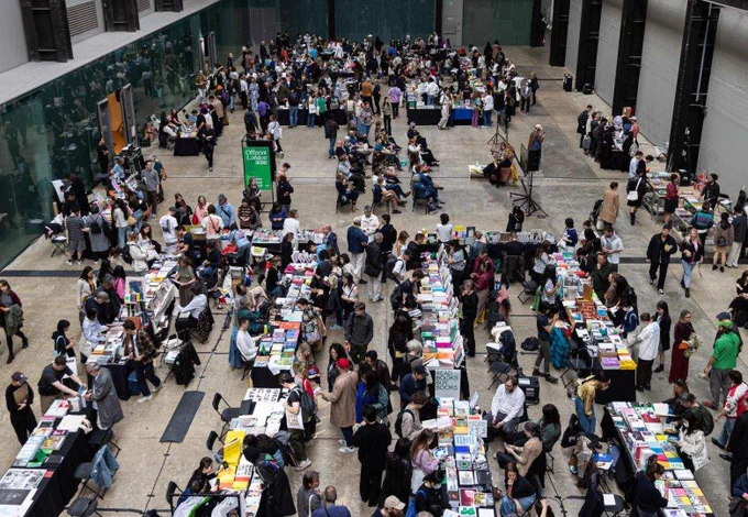 Photograph of a fair in a large hall with lines of tables covered in books and people walking around and surrounding them.
